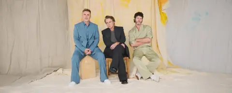 Gary Barlow, Howard Donald et Mark Owen, de Take That, assis dans un décor clair
